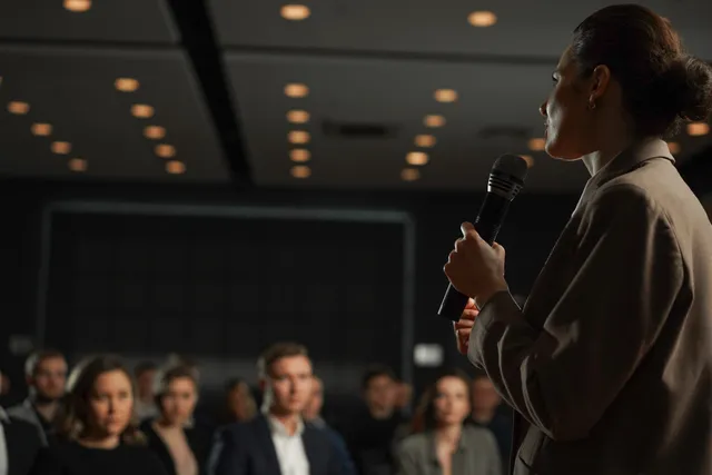 A woman in a tan blazer holds a microphone and addresses an audience in a dimly lit conference hall, with warm ceiling lights overhead and attendees seated in soft focus in the foreground.