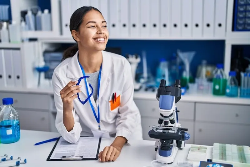 Smiling female scientist in a white lab coat holding a stethoscope at a laboratory workbench with a microscope and scientific equipment.