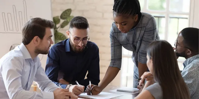 Diverse team of five professionals reviewing documents and collaborating around a table in a modern office with white brick walls.