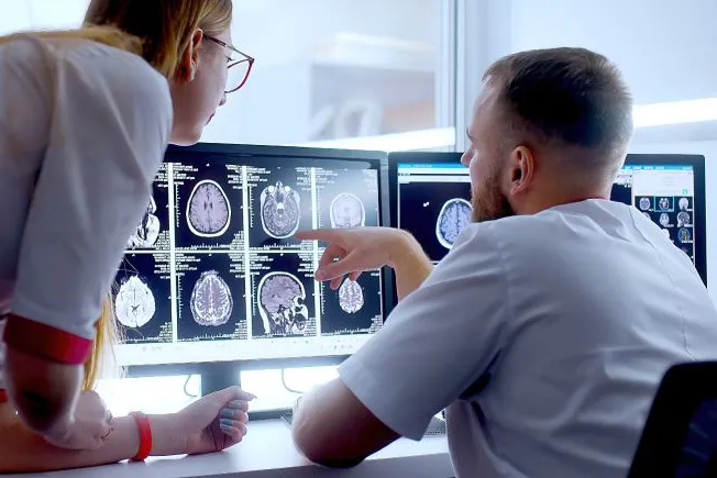 Two medical professionals reviewing brain scan images on dual monitors in a clinical setting.