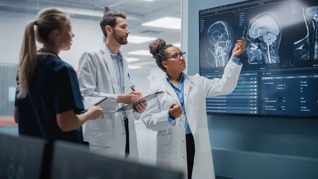 Three medical professionals in lab coats review brain scan images displayed on a large wall monitor in a clinical research setting.