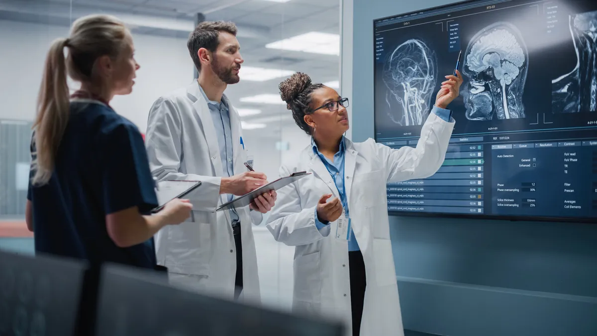 Three medical professionals in lab coats review brain scan images displayed on a large wall monitor in a clinical research setting.