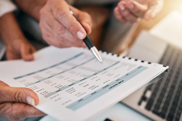 Close-up of a hand using a pen to point at a row in a printed financial spreadsheet held by another person, with an open laptop visible on the desk beside them.