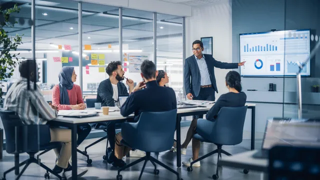 A presenter in a navy blazer points to colorful charts on a wall display while a diverse group of seated colleagues listens around a conference table.