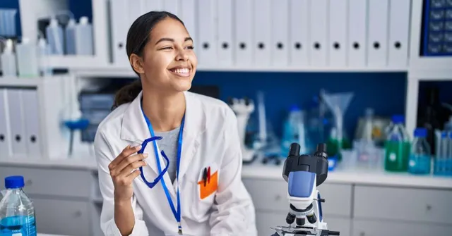 Smiling woman in a white lab coat holding a stethoscope at a laboratory workstation with a microscope and scientific equipment.