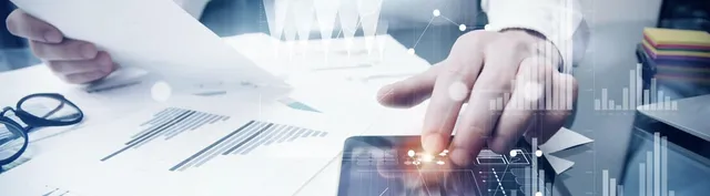 Person pointing at a tablet displaying data visualizations, with printed charts and a magnifying glass on the desk.