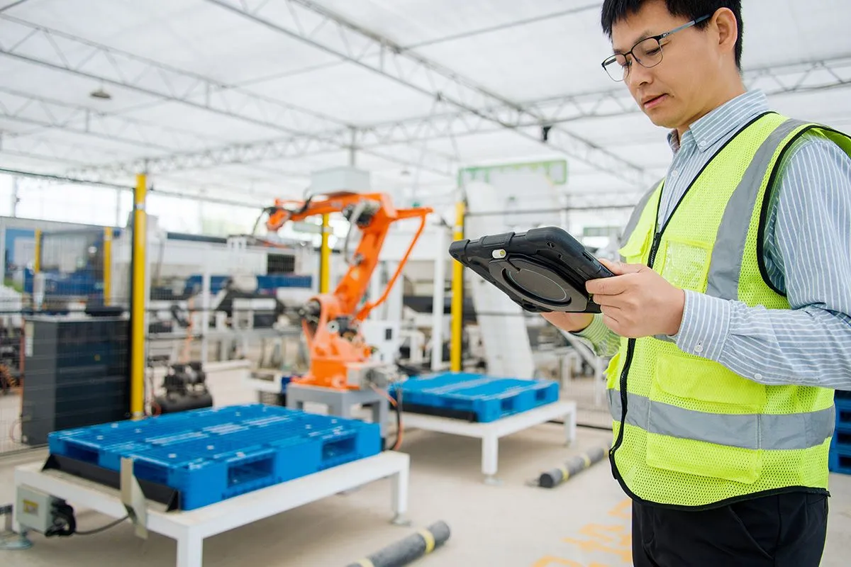 Worker in a safety vest using a tablet on a modern automated manufacturing factory floor with a robotic arm in the background.
