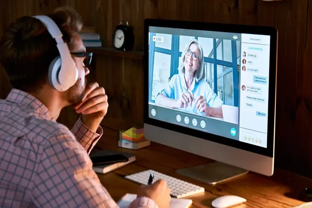 A man wearing white headphones takes notes at a wooden desk while a presenter speaks during a video call on his desktop monitor.