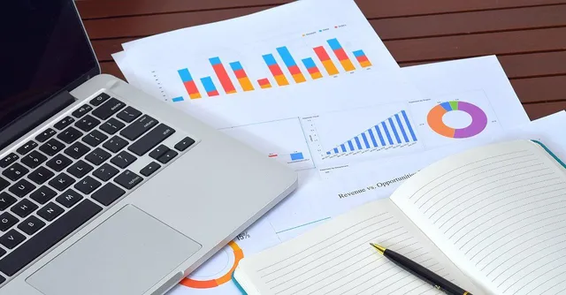 Desk with a laptop and printed data analytics documents including bar charts and a donut chart, with an open notebook and pen.