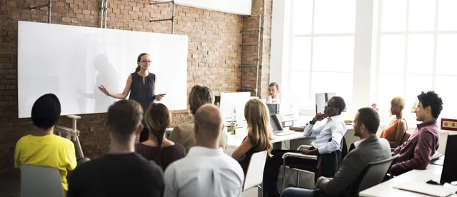 Woman presenting to a group of colleagues in a modern office with exposed brick walls and large windows.