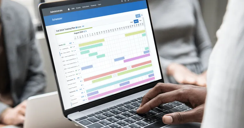 Administrate Scheduler interface showing a Training Plan Gantt chart, with a hand resting on a laptop keyboard.