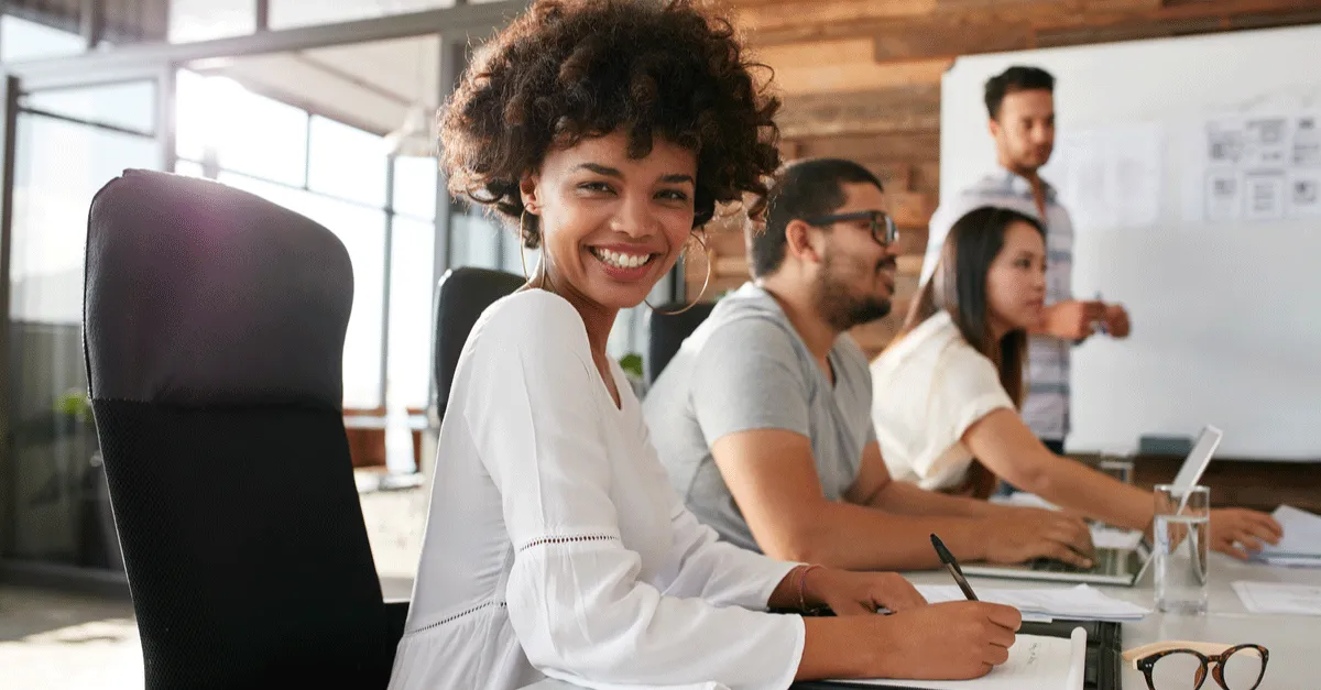 Young professional with curly hair smiling at the camera during a team meeting in a modern office with a whiteboard in the background.