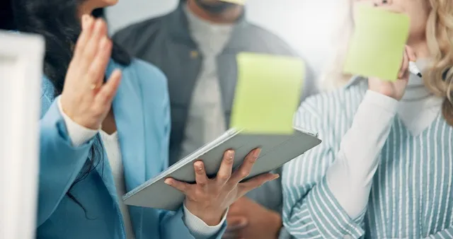 A group of people having a discussion, one person holding a tablet and another writing on a sticky note. The focus is on their hands and device.