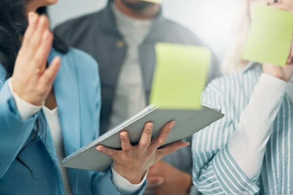 A group of people having a discussion, one person holding a tablet and another writing on a sticky note. The focus is on their hands and device.