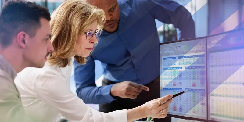 Three professionals focus intently on a computer monitor displaying complex spreadsheets, as one person points at the screen with a pen.