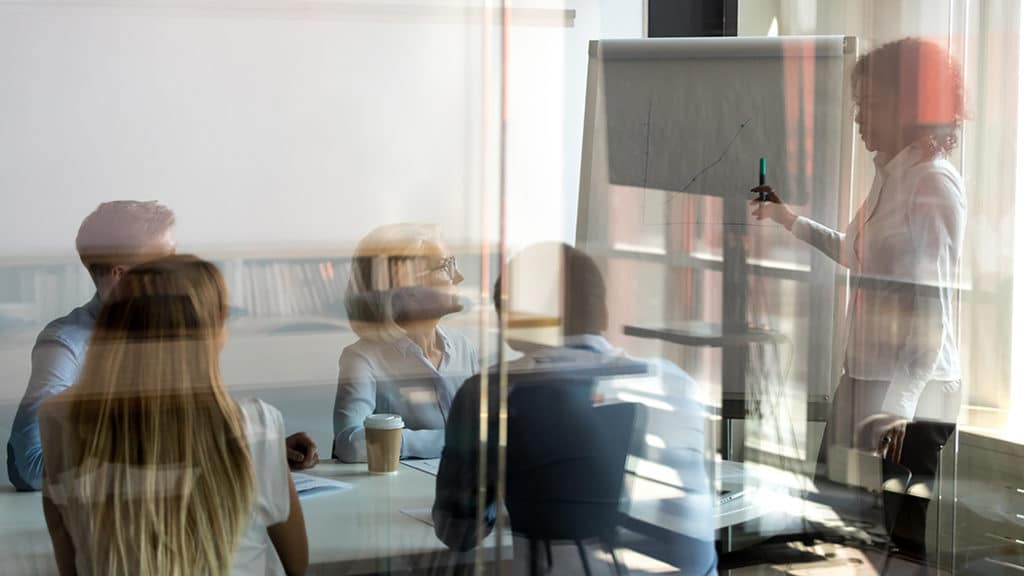 Woman pointing at a chart drawn onto a large pad while presenting findings to a room full of her colleages.