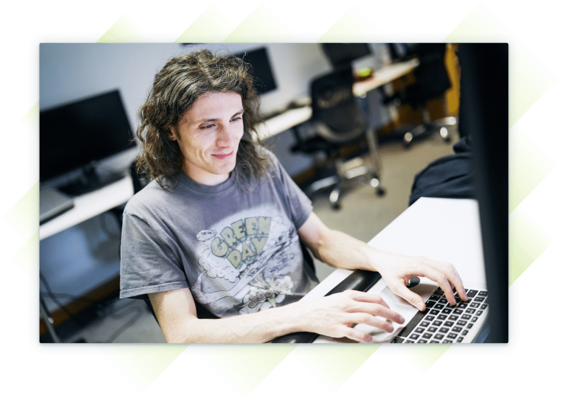 A team member wearing a t-shirt, smiling while working on a laptop at a desk.