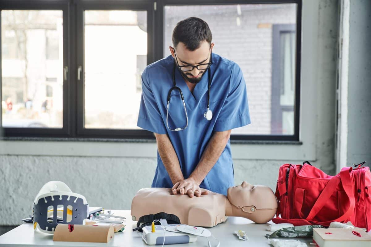 Young paramedic in blue uniform and eyeglasses practicing chest compressions on CPR manikin near defibrillator and first aid kit during medical seminar, life-saving skills development.