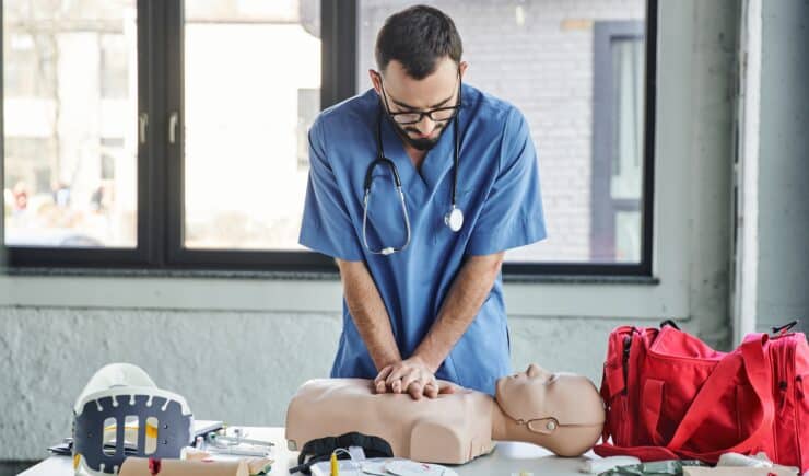 Young paramedic in blue uniform and eyeglasses practicing chest compressions on CPR manikin near defibrillator and first aid kit during medical seminar, life-saving skills development.