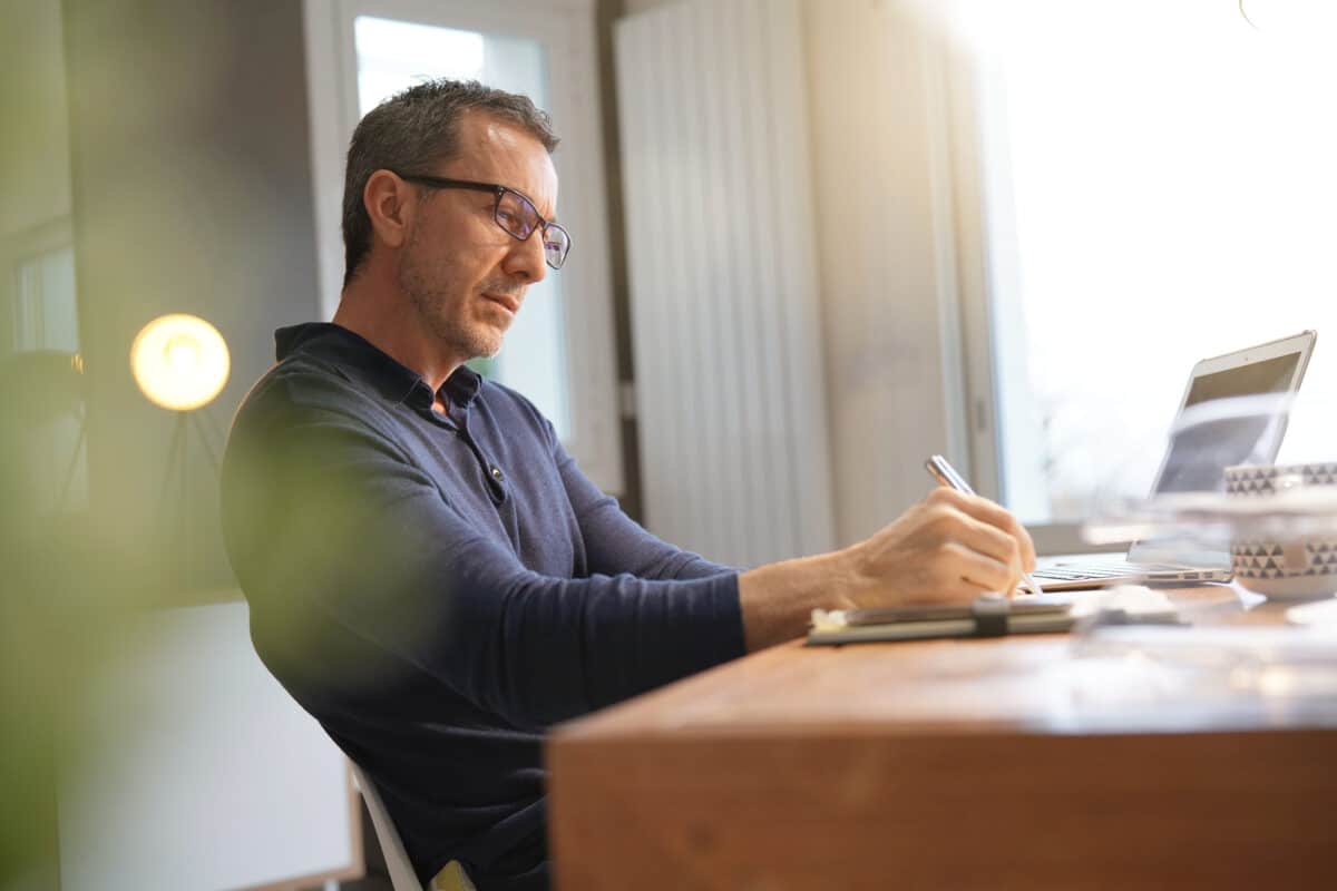 Middle-aged professional taking notes in his office while looking at his laptop in preparation for a meeting.