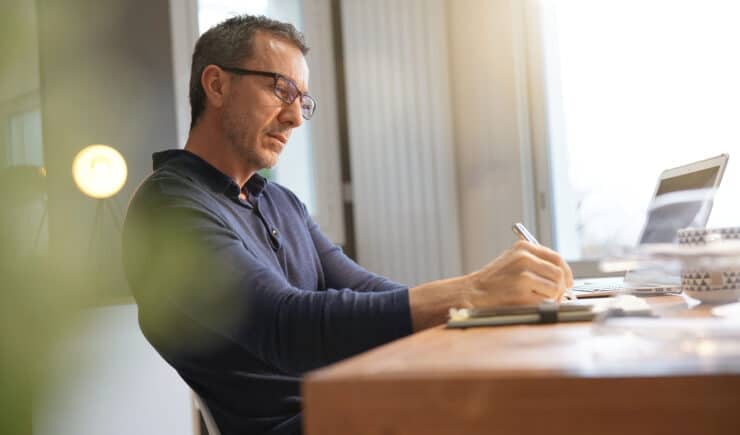 Middle-aged professional taking notes in his office while looking at his laptop in preparation for a meeting.