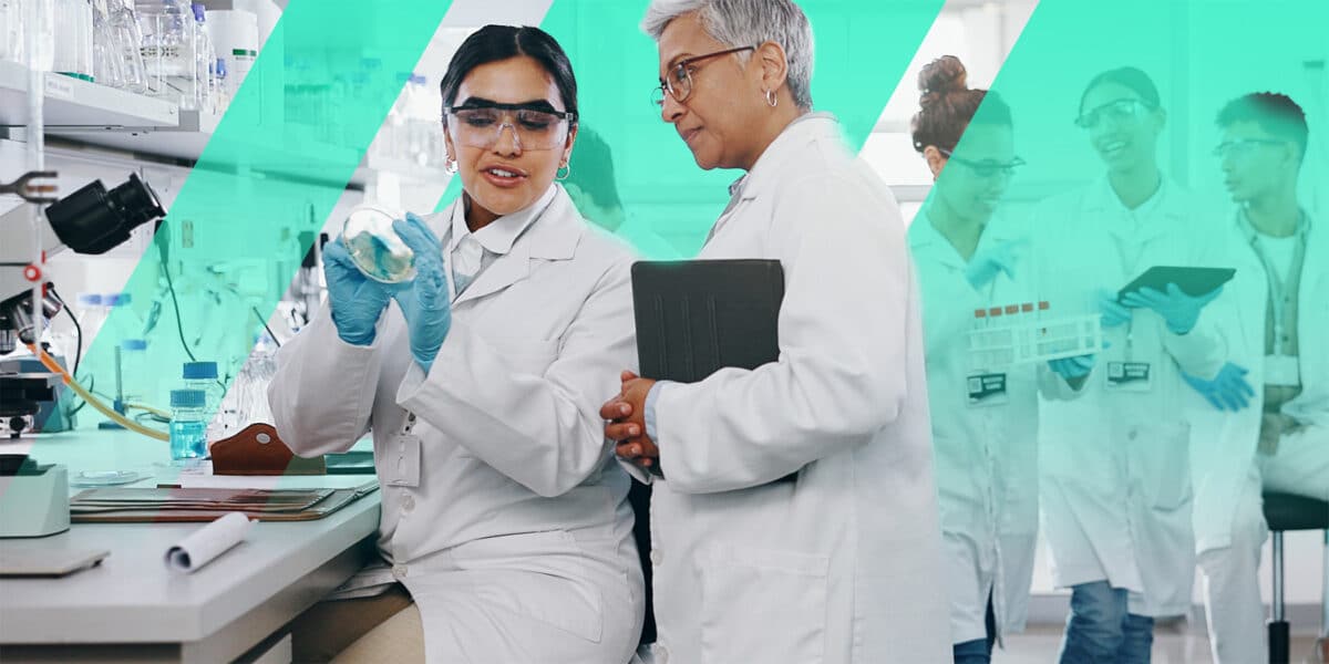 An experienced medical instructor in a white lab coat holds a tablet while guiding a young trainee examining a petri dish in a laboratory setting. This represents instructor-led training in a scientific or medical environment.