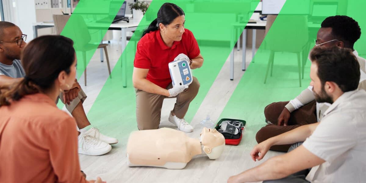 A CPR instructor wearing a red polo shirt and gloves demonstrates how to use an automated external defibrillator (AED) to a group of learners seated in a circle. A CPR training mannequin lies on the floor, along with a medical kit. The setting emphasizes hands-on, instructor-led training for life-saving emergency response.